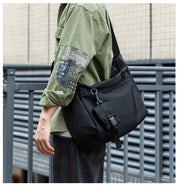 Person wearing a green shirt with patch details and carrying a black shoulder bag against a metallic fence background.