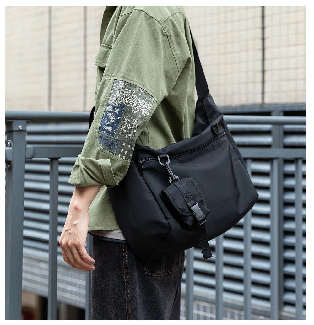 Person wearing a green shirt with patch details and carrying a black shoulder bag against a metallic fence background.
