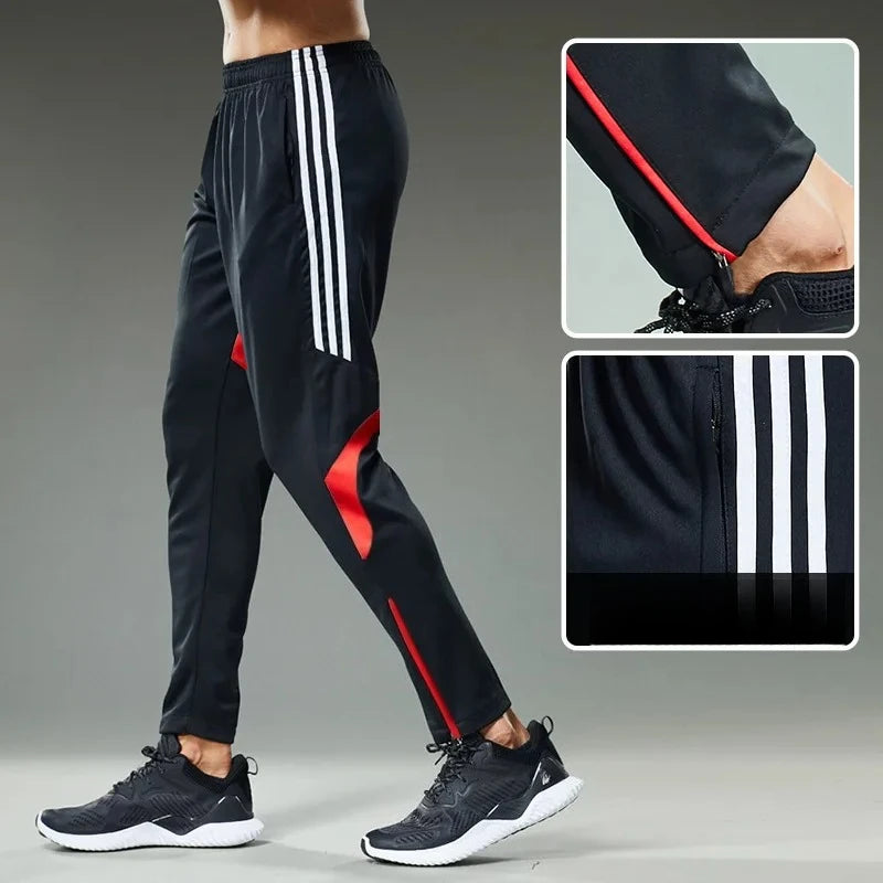 Black tracksuit pants with red accents and white stripes, worn by a person on a gray background.
