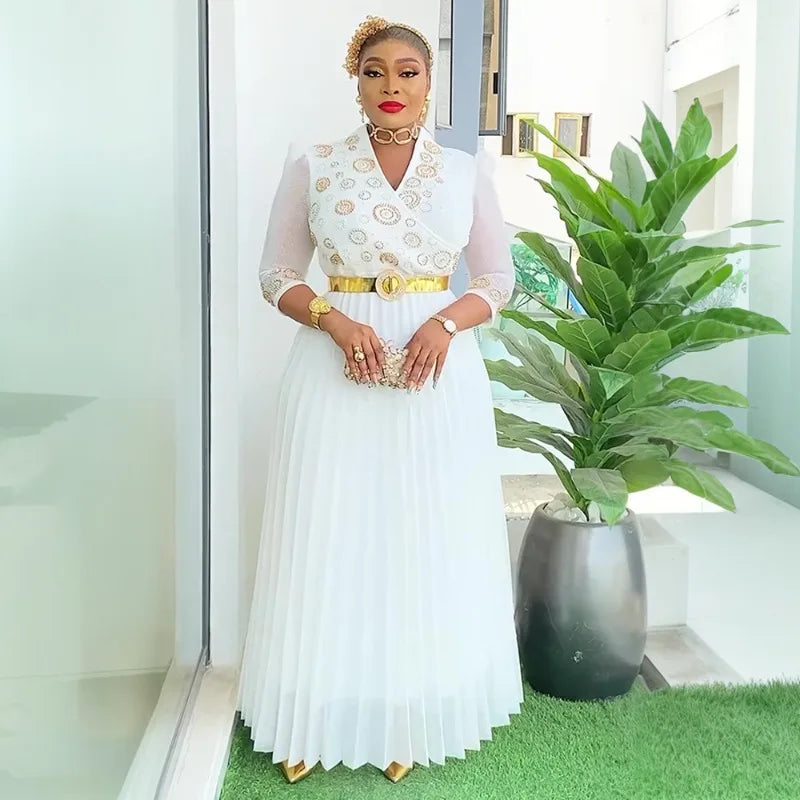 Woman in a white dress with gold accents standing in a modern indoor setting with a plant.