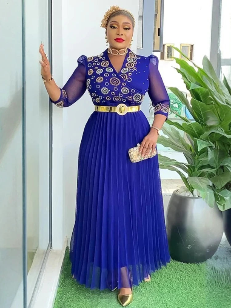 Woman in a blue dress with gold accents standing indoors next to a plant.