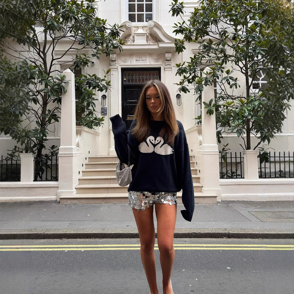 Woman wearing a navy blue sweater with a white swan design, standing on a street in front of a white building.